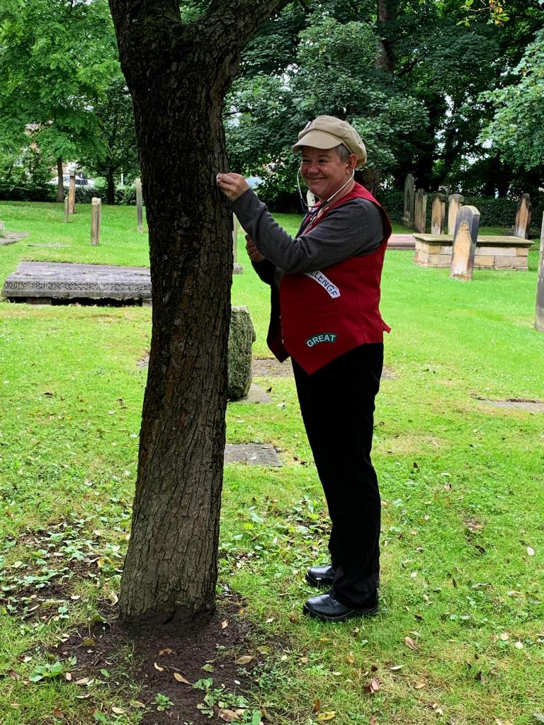 A small white woman with cropped hair under a beige peaked cap, wearing a uniform-like outfit of black trousers and a red waistcoat patched with words like 'challenge' and 'great'. She is standing next to a tress in a churchyard, you can see well-maintained grass and headstones behind her. She is using a stethoscope to listen to the tree, and is laughing.