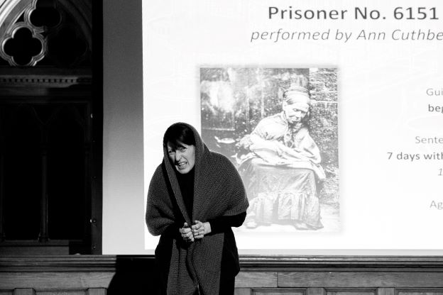 Ann, a white woman with straight dark hair, is hunched over with a roughly-woven shawl over her head and shoulders, adopting the persona of a much older and more crone-like woman. We can see a projection on a screen behind her, showing an early photograph of an old woman sitting crouched against the walls of Holloway Prison.