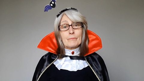 An older woman with straight grey-white hair and glasses is looking serious but is wearing a joke-shop black satin Dracula cape with a high red collar, and a headband with a cartoon bat on a wire above her head.