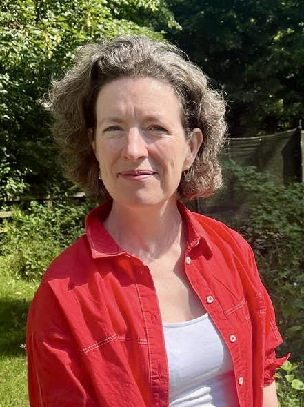 White woman, forties, short brown wavy hair, intelligent direct and enquiring expressions, wearing red shirt open over white vest, in a sunny garden
