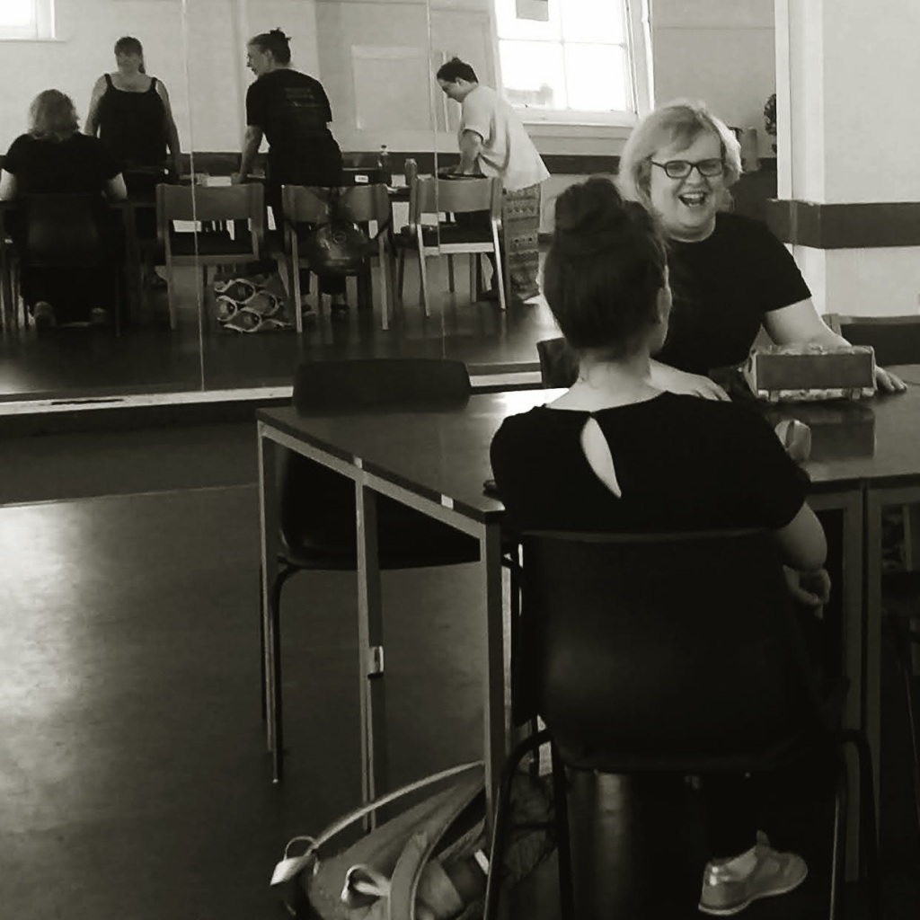 Black and white image of a large rehearsal room with lots of natural light. Two women sit at a table, we can see one from the back (young, long dark hair up in a bun) and one facing us laughing (white woman in glasses, short blonde hair). In the large wall mirrors beyond we can see three other woman standing, talking and taking things out of bags.