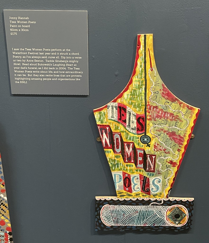 A gallery display. A painted board cut in the shape of a pen nib has colourful illustration on it saying 'Tees Women Poets' and 'words sewn together form stories'