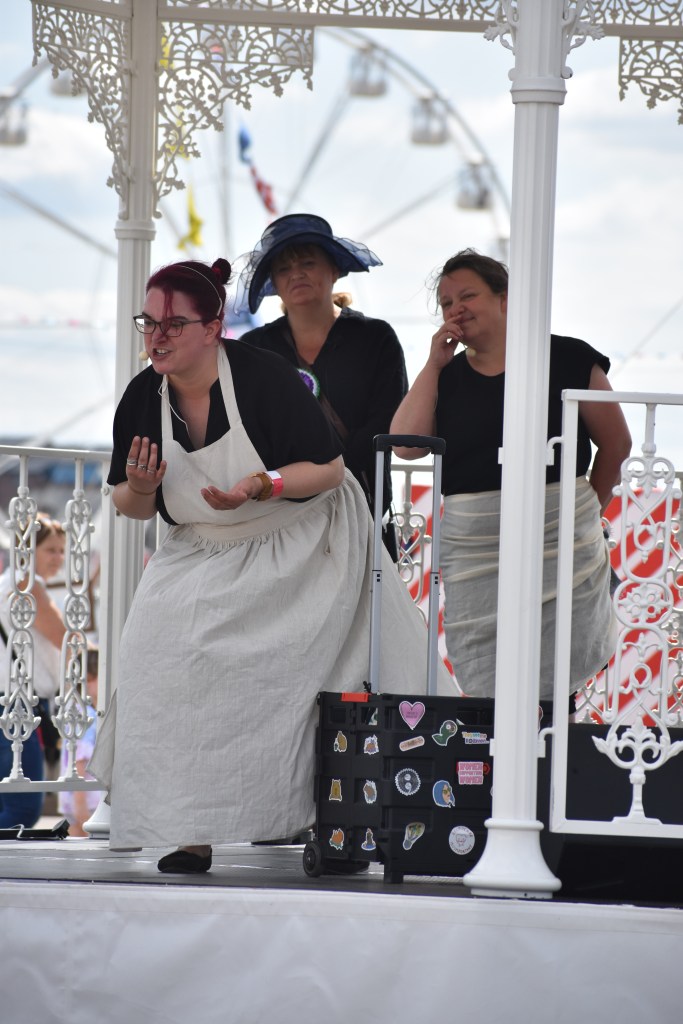White woman in a white fishmonger's apron over a black dress gestures at the audience - two other women in the background hold their noses as if smelling something bad