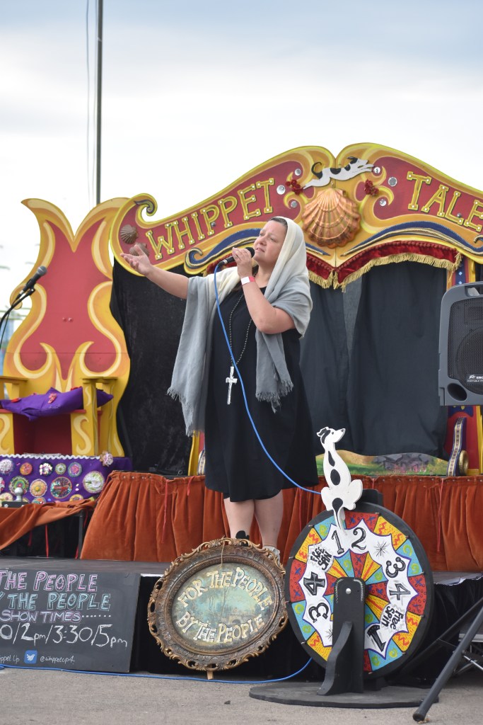 white woman draped in a grey shawl-scarf, wearing a black dress with a large fake crucifix is gesturing and trying to look holy