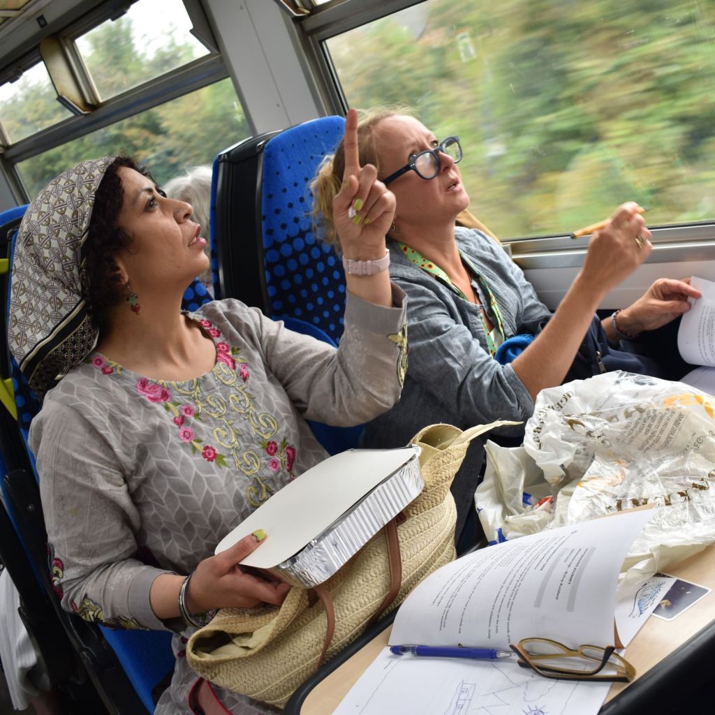 Two women sit next to each other at a train table, looking up and pointing outside. One 