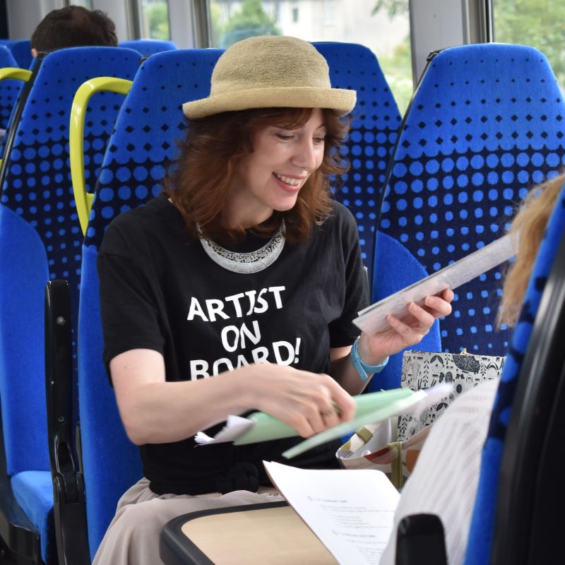Carmen, a white woman in her thirties, is sitting on a train organising a bag full of papers. She is wearing a jaunty straw summer hat and a t shirt that says Artist On Board.