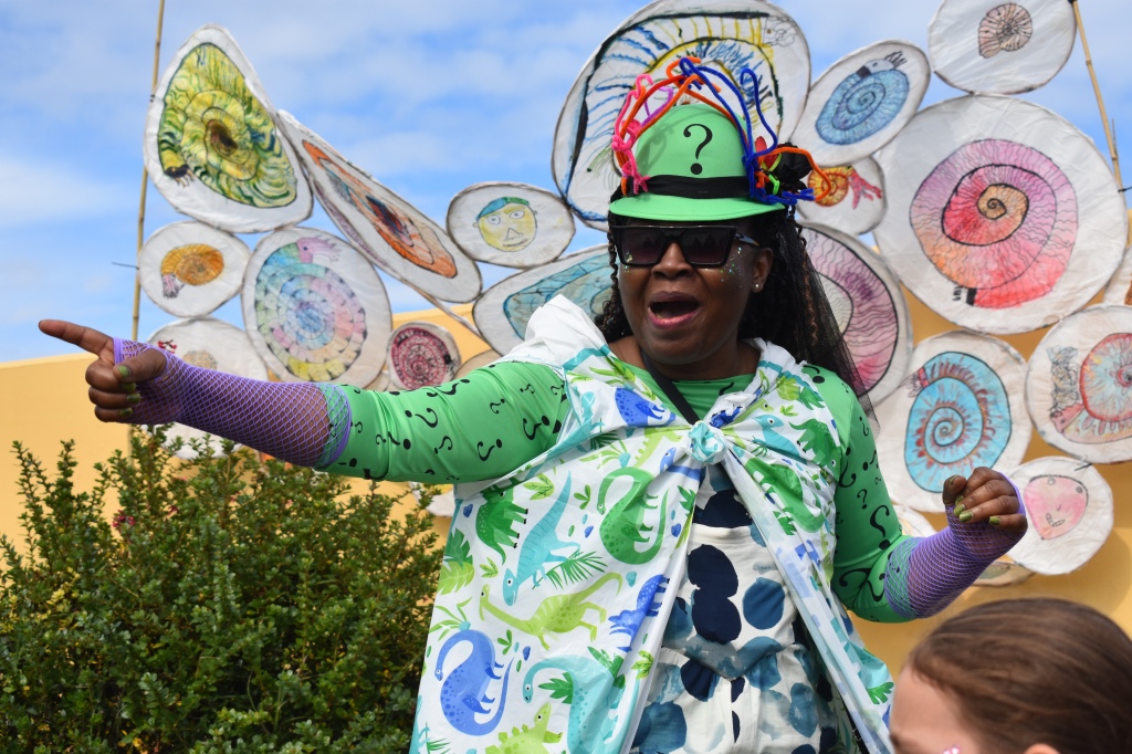 A black woman points and gestures in front of an outdoor installation of withy-and-tissue ammonites drawn by kids. The women wears a green hat and a green top covered in question marks, and purple fishnet gloves.
