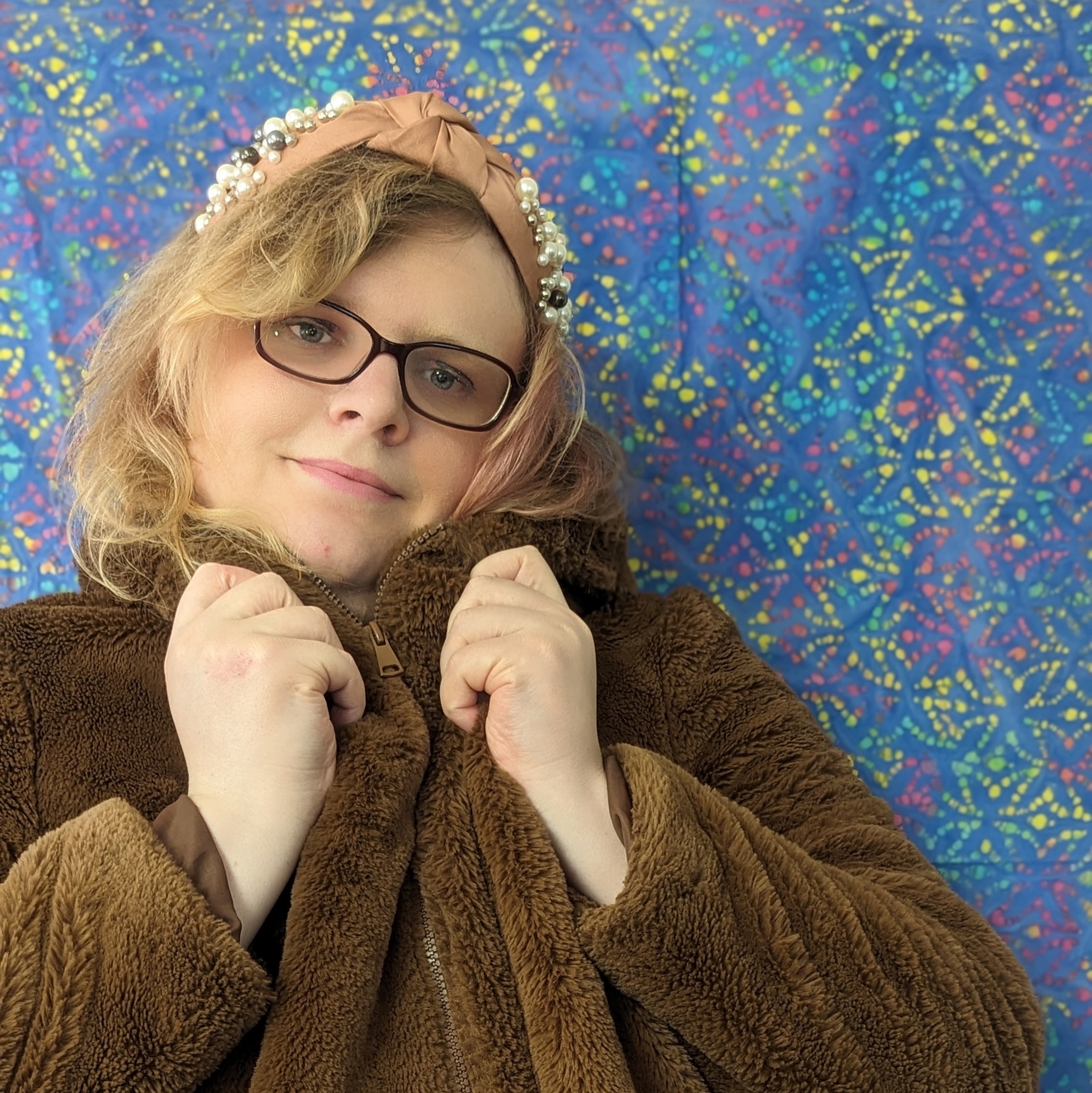 A young white woman with blonde hair stands in front of a bright blue patterned batik hanging. She has a pearl and knotted beige silk headband on and is holding the collar of her fake fur coat up to her chin