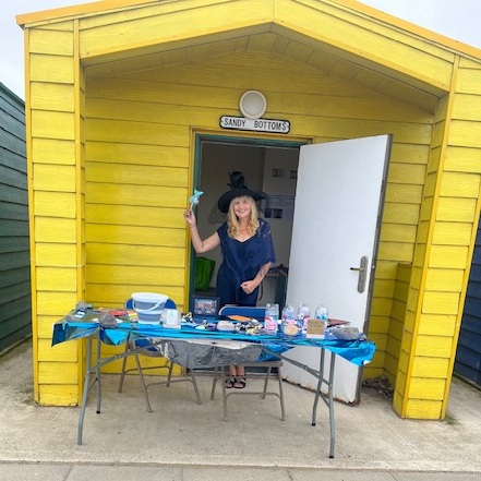 Danne, a white woman in her fifties with long blonde hair, is dressed as a witch and sitting in the entrance of a yellow beach hut called 'Sandy Bottoms'