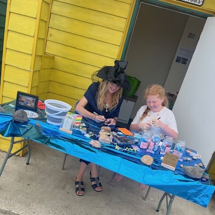 Dianne in her witches hat is sitting with a young girl at a shiny blue table covered with sea shells, paper, and other crafting materials