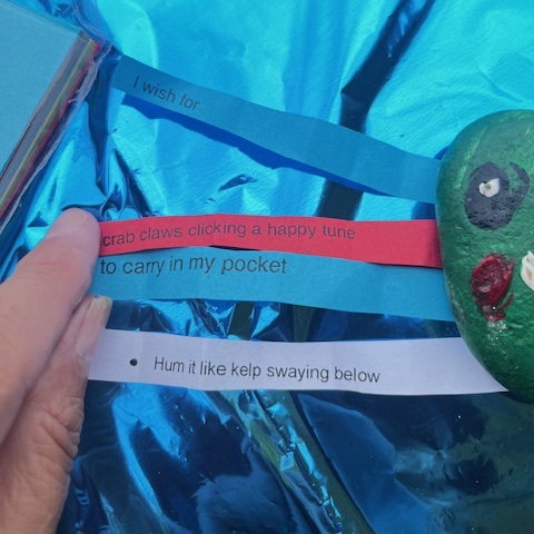 close up of three strips of paper against a shiny blue table; the strips have lines of poetry on them - I wish for crab claws clicking a happy tune to carry in my pocket